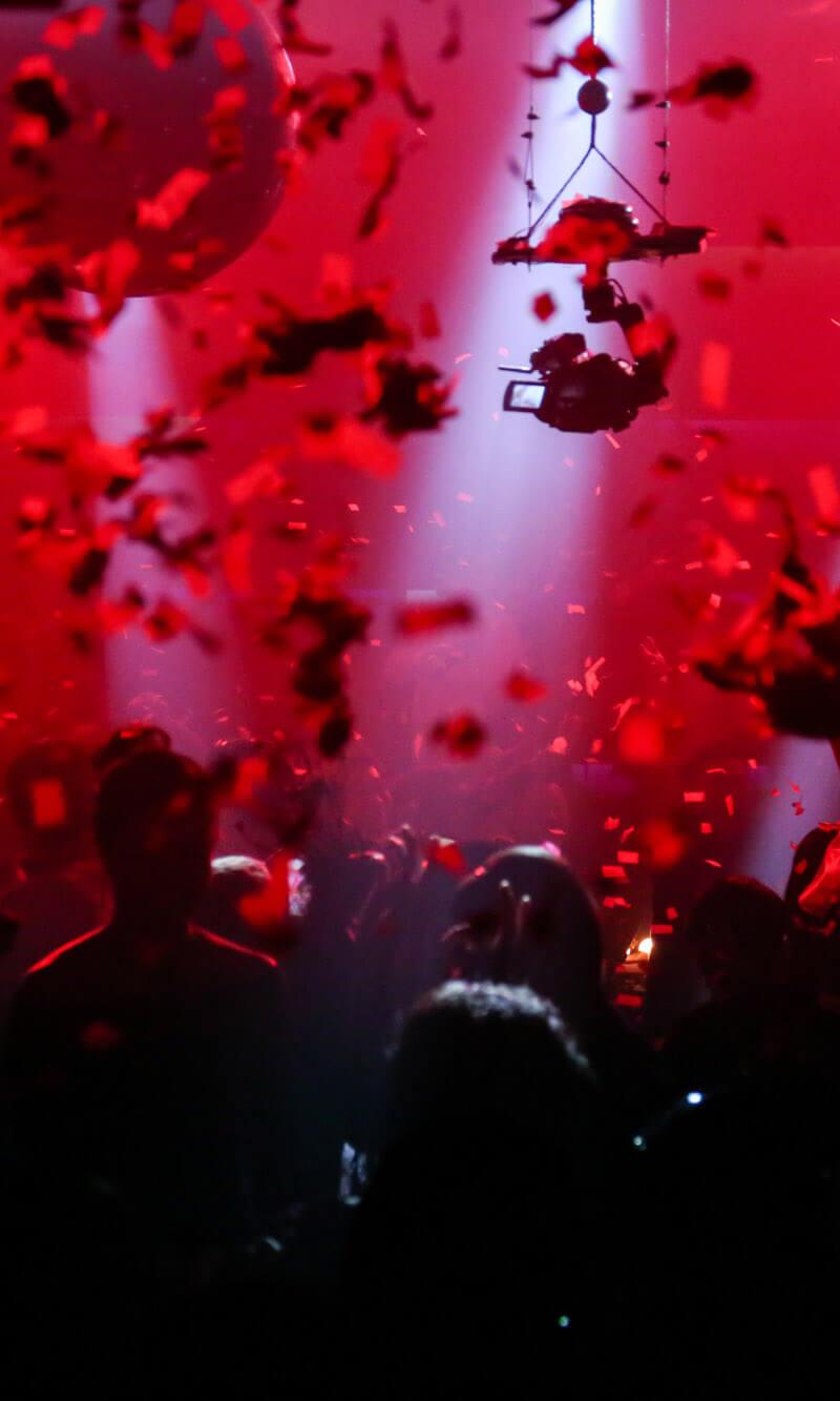Man sieht die Hände einer tanzenden Menschenmenge im rotem Licht. Konfetti ist in der Luft.
