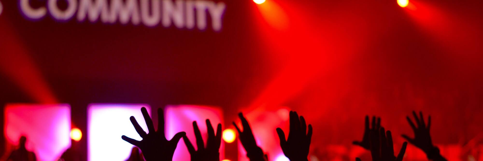 Man sieht die Hände einer feiernden Menschenmenge im rotem Licht. Das Wort 'Community' wird verschwommen an die Wand darüber projiziert.
