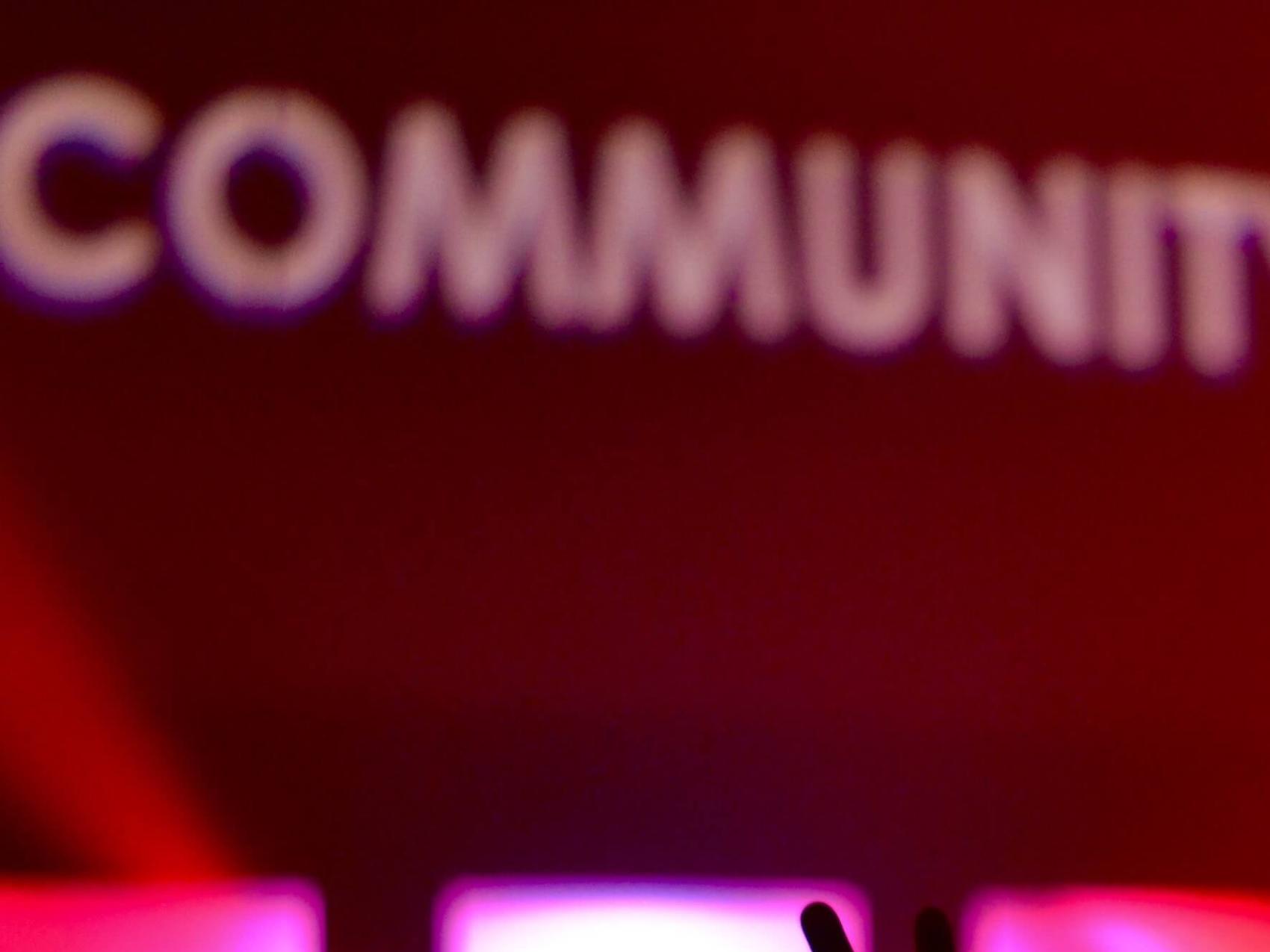 Man sieht die Hände einer feiernden Menschenmenge im rotem Licht. Das Wort 'Community' wird verschwommen an die Wand darüber projiziert.